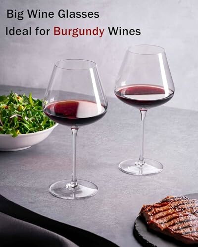 Two big wine glasses with red wine, salad, and steak.