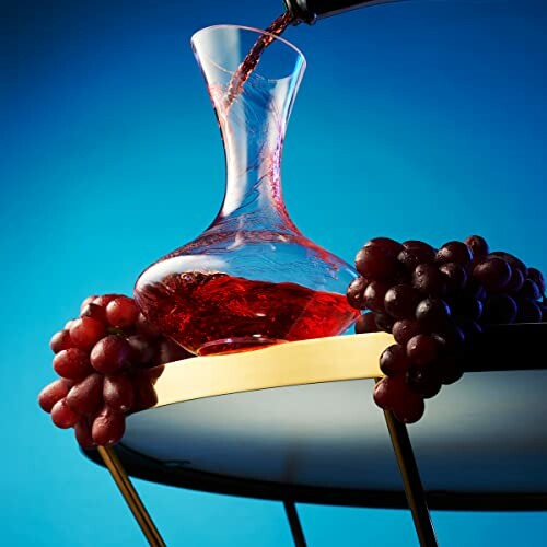 Red wine being poured into a decanter with grapes on a table.